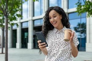 sorridente hispânico empresária desfrutando uma café pausa ao ar livre, usando dela Smartphone com a escritório construção pano de fundo. foto