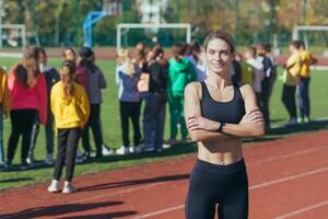 uma fêmea fisica Educação professor às escola sorrisos e parece às a Câmera, a atleta treinador em a fundo do crianças, retrato. foto