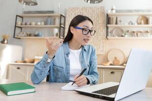 jovem ásia aluna estudando às casa remotamente, mulher assistindo conectados curso sentado dentro cozinha usando computador portátil e bloco de anotações foto