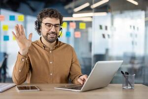 alegre homem de negocios tendo uma chamar, acenando Olá para a Webcam em dele computador portátil dentro uma moderno escritório contexto. foto