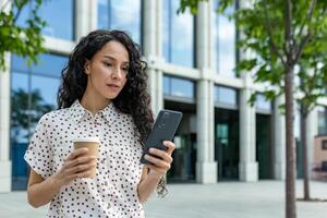 jovem hispânico empresária com encaracolado cabelo desfrutando uma café quebrar, navegando dela Smartphone, lado de fora uma moderno escritório prédio. foto