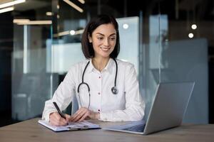 atraente fêmea terapeuta dentro profissional uniforme olhando às computador portátil tela e fazer notas de mão em prancheta. contemporâneo hospital trabalhador fornecendo conectados Serviços e consultas para cliente. foto