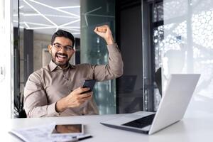 bem sucedido homem de negocios vencedora dentro escritório às ambiente de trabalho, homem sorridente e olhando às Câmera recebido conectados ganhar notificação ganhar, empregado às mesa com computador portátil a comemorar triunfo. foto