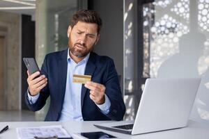 sério jovem masculino homem de negocios sentado dentro escritório às escrivaninha com computador portátil e preocupado olhando às crédito cartão, segurando telefone, tendo financeiro e técnico dificuldades. foto