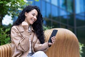 jovem mulher vencedora recebido bem sucedido conectados mensagem em telefone, empresária feliz a comemorar sentado em Banco lado de fora escritório prédio, usando aplicativo em Smartphone. foto