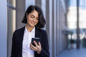 uma jovem lindo o negócio mulher é caminhando lado de fora a escritório prédio, uma mulher dentro o negócio roupas é sorridente, usando a inscrição em a telefone, navegando a Internet, digitando uma mensagem. foto