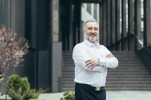 retrato do uma Senior homem, uma professor, uma conferencista em pé dentro uma branco camisa contra a fundo do uma moderno construção do a educacional instituição. sorrindo, Veja às a Câmera foto