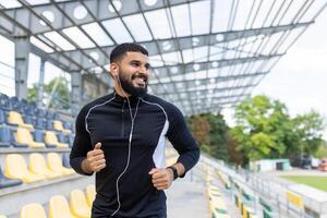 uma alegre jovem homem corrida dentro uma Esportes estádio, desfrutando uma exercite-se com música através dele fones de ouvido. foto
