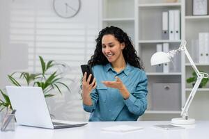 uma sorridente, profissional mulher envolve dentro uma chamar. ela é sentado às uma organizado, organizado escritório mesa, gesticulando enquanto olhando às dela Smartphone. foto