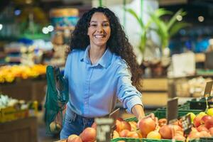 retrato do feliz mulher comprador dentro supermercado, hispânico mulher escolhe maçãs e frutas sorridente e olhando às Câmera, com mercearia cesta escolhe bens foto