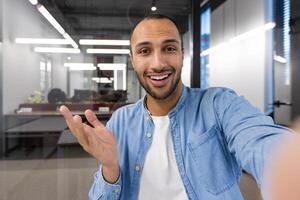 uma alegre jovem homem capturando uma selfie com uma Smartphone dentro uma bem iluminado contemporâneo escritório contexto, expressando positividade e confiança. foto