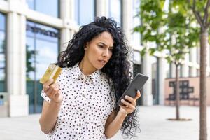 chateado e desapontado mulher recebido dinheiro transferir erro, hispânico mulher com banco crédito cartão e telefone enganado caminhando baixa rua lado de fora escritório prédio. foto