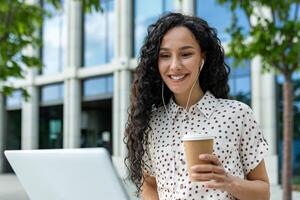 uma jovem profissional mulher leva uma relaxante café pausa ao ar livre, usando dela computador portátil contra a pano de fundo do uma moderno escritório prédio. isto sincero momento captura a Saldo entre trabalhos foto