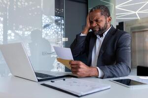 nervoso e chateado homem de negocios dentro escritório às local de trabalho recebido enviar envelope notificação mensagem com mau notícia falência, africano americano homem triste e pensativo lendo documento. foto