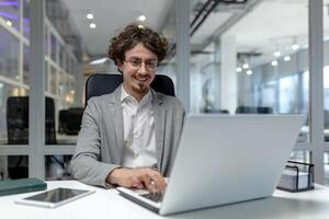 jovem, profissional masculino escritório trabalhador com encaracolado cabelo digitando em uma computador portátil, exibindo eficiência e uma contente comportamento no meio a corporativo labuta. foto