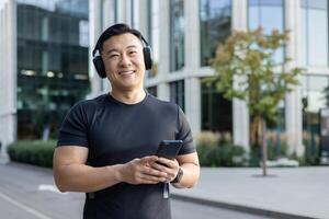 retrato do uma jovem ásia homem em pé em uma cidade rua vestindo fones de ouvido e ouvindo para música em a telefone. tocam Esportes, corre, sorrisos e parece às a Câmera. foto