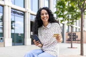 retrato jovem lindo mulher com encaracolado cabelo, sentado em uma Banco lado de fora a escritório prédio, sorridente e olhando às Câmera, segurando uma telefone, banco crédito cartão, encomenda conectados Serviços, compras. foto