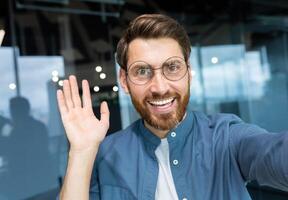 maduro homem dentro escritório vestindo camisa e óculos sorridente e olhando às Smartphone Câmera, homem de negocios levando selfie e falando em ligar usando Smartphone, programador acenando mão cumprimento gesto. foto