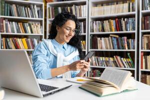 feliz hispânico latino aluna usando dela Smartphone dentro uma biblioteca. Educação, tecnologia e Aprendendo conceito. foto