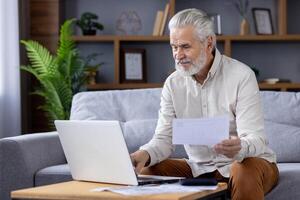 idosos de cabelos brancos homem dentro uma casual camisa parece às papelada enquanto sentado em uma sofá, usando uma computador portátil dentro uma bem mobiliado vivo sala. foto