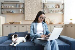 jovem lindo ásia trabalhador autonomo trabalhando a partir de casa remotamente, mulher sentado em sofá com animal pequeno cachorro, jack russell terrier raça, usando computador portátil foto