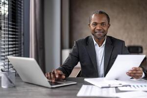 profissional africano americano homem de negocios analisando papelada com computador portátil e tábua em dele escrivaninha dentro uma moderno escritório contexto. foto