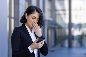 empresária recebido notificação mensagem com mau notícias, mulher lendo conectados em telefone, chateado e confuso triste usando Smartphone aplicativo, lado de fora escritório prédio, caminhando dentro o negócio terno. foto