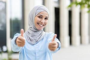 uma alegre, jovem mulher vestindo uma hijab dá uma Duplo polegares acima com uma largo, confiante sorriso, sinalização aprovação e sucesso. foto