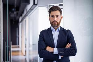 retrato do uma confiante profissional masculino executivo com uma barba em pé dentro uma moderno escritório meio Ambiente com dele braços cruzado. foto