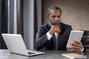 a africano americano homem de negocios dentro uma terno trabalhando atentamente com uma tábua e computador portátil às dele escritório escrivaninha. uma representação do moderno multitarefa e tecnologia usar dentro negócios. foto