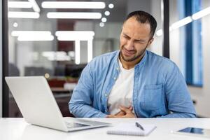 indiano homem de negocios experimentando estômago dor enquanto trabalhando em uma computador portátil dentro uma moderno escritório contexto, aparecendo angustiado. foto