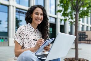 a independente jovem mulher dentro casual o negócio vestuário é focado em dela trabalhos em uma computador portátil enquanto sentado lado de fora em uma brilhante, ensolarado dia. foto