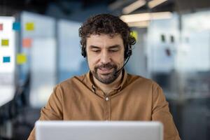 focado hispânico homem usando uma fone de ouvido e trabalhando diligentemente em dele computador portátil às uma moderno escritório área de trabalho. foto