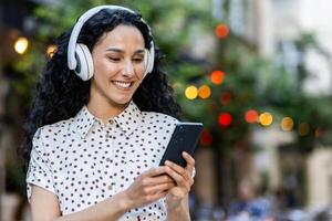 jovem lindo hispânico mulher anda em tarde cidade dentro fones de ouvido, aluna escuta para música on-line, usa a inscrição em uma Smartphone, detém a telefone dentro dela mãos, lê mensagens, sorri. foto