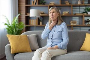 doente mulher sentado em sofá com amarelo almofadas e sofrimento a partir de dolorido garganta enquanto segurando mão em pescoço. pouco saudável fêmea tendo sintomas do gripe ou laringite doença enquanto gastos dia às às lar. foto