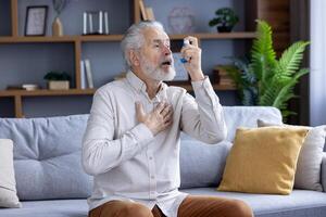 idosos caucasiano homem experimentando a asma ataque, usando uma azul inalador enquanto sentado em uma sofá dentro uma moderno vivo sala. foto