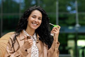 uma alegre hispânico mulher ao ar livre usando uma remédio spray para garganta tratamento, expressando satisfação com uma polegares acima. foto