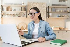 Aprendendo on-line. uma jovem menina aluna leva a exame remotamente através uma ligar com uma professor. ele senta às casa dentro a cozinha com uma computador portátil, parece às a Câmera, estudos, escreve dentro uma caderno. foto