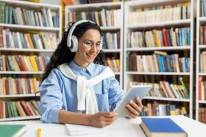 uma alegre jovem aluna envolve dentro conectados Aprendendo com tábua e fones de ouvido no meio uma da biblioteca estantes. foto