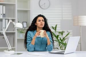 uma mulher dentro uma azul camisa goza uma café pausa às dela limpar, branco escritório espaço com uma computador portátil e interior plantas. foto