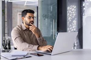 homem pensando dentro escritório às local de trabalho com computador portátil, homem de negocios preparando financeiro relatório, digitando em teclado, financista contador dentro óculos e camisa segurando mão para queixo. foto