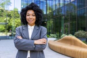 confiante adulto empreendedor dentro roupa formal em pé com guardada mãos em cintura em fundo do moderno prédio. africano americano mulher pronto para Novo o negócio dia e incorporando Liderança e sucesso. foto