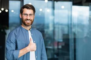 retrato do homem de negocios dentro casual camisa, homem com barba e óculos sorridente e olhando às Câmera mostrando polegares acima afirmativo, o negócio proprietário trabalhando dentro escritório. foto