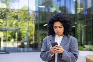 chateado jovem africano americano empresária em pé lado de fora escritório construção e olhando preocupado às telefone tela. fechar-se foto. foto