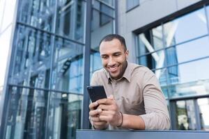 bem sucedido homem de negocios lado de fora moderno escritório construção usando Smartphone, homem lendo mensagem e navegando rede Páginas, africano americano homem dentro camisa segurando telefone digitando mensagem. foto