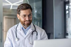 fechar-se foto do uma jovem sorridente masculino médico sentado dentro a escritório e falando em uma ligar em uma computador portátil.