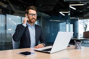 retrato do uma bem sucedido homem de negocios dentro uma moderno escritório, uma homem dentro uma o negócio terno usa fones de ouvido para uma ligar e controlo remoto comunicação, a investidor parece às a Câmera e usa uma computador portátil. foto