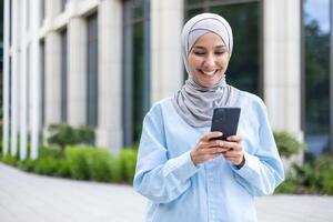 uma jovem lindo o negócio mulher dentro uma hijab anda em através a cidade, uma muçulmano mulher detém uma telefone dentro dela mãos, usa a inscrição em uma Smartphone, sorrisos, navega a Internet. foto