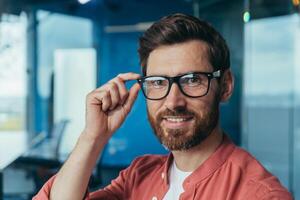 fechar-se foto do jovem bem sucedido arquiteto com barba, maduro masculino desenhador olhando às Câmera e sorridente dentro óculos e vermelho camisa perto janela dentro escritório.