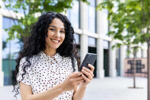 alegre mulher com encaracolado cabelo, usando Smartphone ao ar livre dentro urbano contexto, exalando positividade e moderno estilo de vida. foto